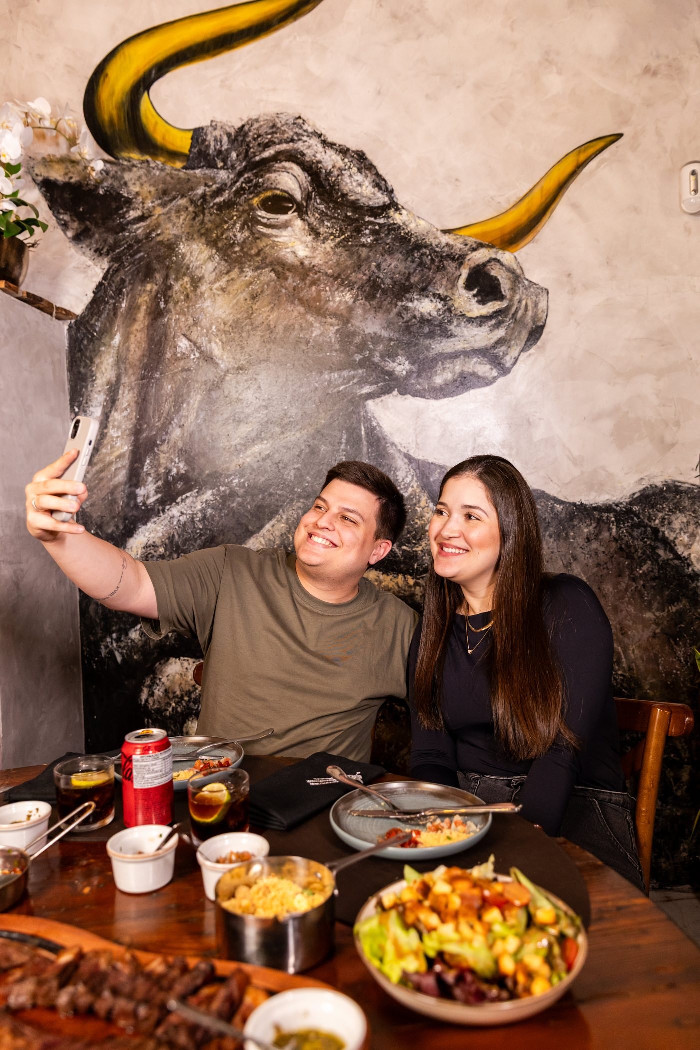 Casal fazendo selfie em frente ao mural do touro, mesa posta com cortes e guarnições